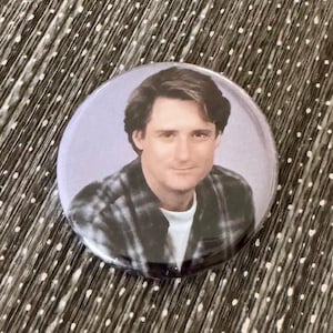 May include: A round button pin featuring a man in a plaid shirt and white undershirt. The pin has a light purple background and a glossy finish. The pin is approximately 2 inches in diameter.