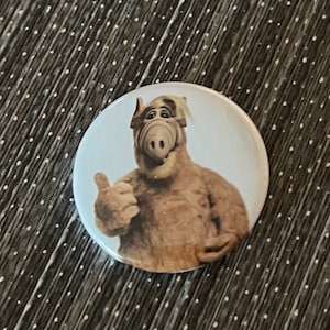 May include: A round button featuring a brown and white alien character with a large nose and a thumbs up gesture. The character is on a white background.