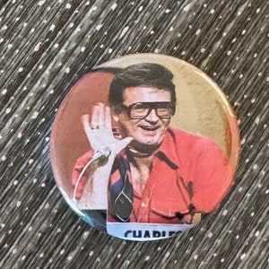 May include: A red, white, and blue button with a photo of a man wearing glasses and a red shirt. The text "CHARLIE" is visible at the bottom of the button.