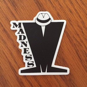 May include: Black and white sticker with a silhouette of a person wearing a fedora and a suit. The word "MADNESS" is written vertically on the left side of the sticker.