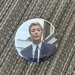May include: A round button featuring a man in a suit and tie. The button has a glossy finish and a white background. The man is looking forward. The button is a collectible item.
