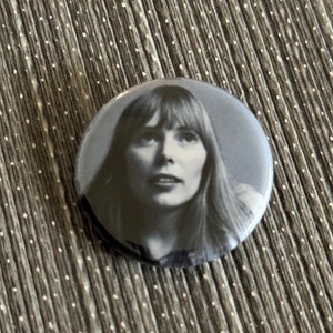 May include: A black and white button featuring a photo of a woman with long hair looking up.