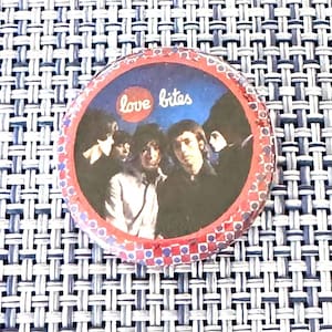 May include: A circular pinback button featuring a dark blue background with a group of people in black and white. The text "love bites" is printed in white inside a red circle. The border has a red and blue checkered pattern.
