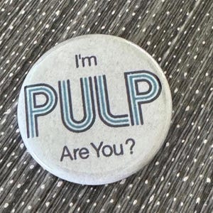 May include: A round button with the text "I'm PULP Are You?" in a teal and blue font. The button has a light gray background and is set against a textured gray surface.