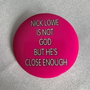 May include: A pink button with white text that reads "Nick Lowe is not God but he's close enough."