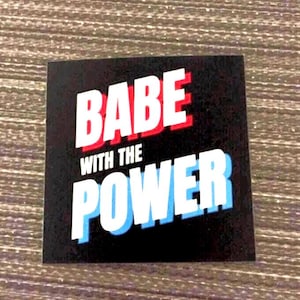 May include: Black and white sticker with the text "BABE WITH THE POWER" in red and blue letters.