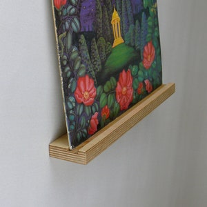 Könnte beinhalten: Ein Vintage-Schallplattenalbum mit dem Titel "Der Goldene Pavillon" wird auf einem Holzregal präsentiert. Das Albumcover zeigt ein Blumendesign mit roten Rosen, Schmetterlingen und einer Landschaftsszene. Das Regal ist aus hellem Holz.