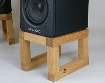 May include: Black studio monitor speakers on wooden stands. The speakers have a black finish and the text "M-AUDIO" and "BX5" printed on the front. The stands are made of light brown wood.