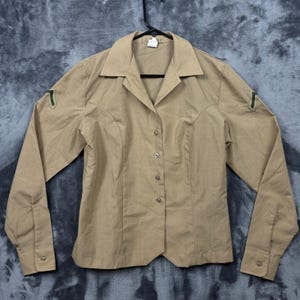 May include: A tan long-sleeved button-down shirt with a pointed collar and a front button closure. The shirt features dark green chevron-style military-inspired patches on both upper sleeves and includes a small white tag at the inner neckline.