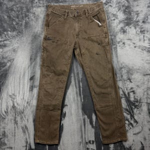 May include: A pair of brown utility pants featuring a button and zipper fly closure, belt loops, and reinforced double-layer fabric panels on the front of the thighs. The pants include side pockets and a hammer loop on the side for utility use.