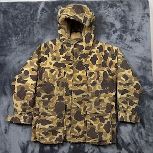 May include: A hooded jacket featuring a brown and tan camouflage pattern. The garment includes a front zipper closure, two chest pockets with flaps, and two large lower pockets with flaps. The sleeves have adjustable cuffs with hook and loop fasteners.