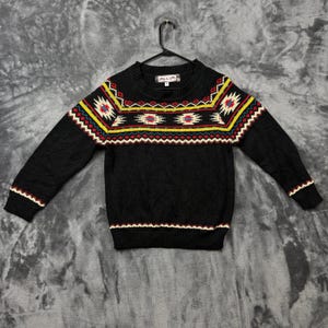 May include: A black long-sleeve sweater featuring a crew neckline and a colorful geometric pattern across the chest and shoulders. The pattern includes red, yellow, white, and blue zig-zag and diamond shapes. A black hanger is attached to the collar.