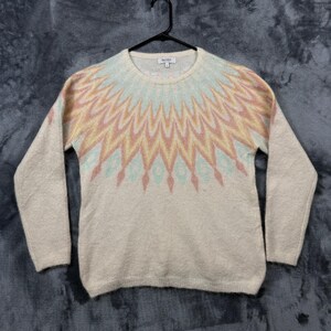 May include: A beige long-sleeve sweater featuring a round neckline and a textured knit finish. The upper chest and shoulder area display a prominent sunburst pattern in soft shades of pastel pink, yellow, and light blue. A tag reads Barneys New York.