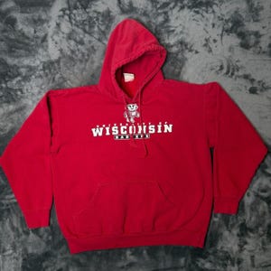 Könnte beinhalten: Ein leuchtend roter Kapuzenpullover mit einer weißen Grafik eines Dachs-Maskottchens über dem Schriftzug UNIVERSITY OF WISCONSIN BADGERS. Der Pullover verfügt über eine Kängurutasche auf der Vorderseite, lange Ärmel und eine Kapuze mit Kordelzug.