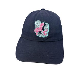 Puede incluir: Una gorra de béisbol azul marino con un diseño bordado en la parte delantera. El bordado muestra el texto "Bury the Past" en una fuente estilizada, con letras blancas y rosas delineadas por una forma de nube azul claro en la corona.
