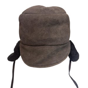 May include: A dark brown suede hat featuring a rounded crown and a wide brim that curves slightly upward. The hat includes black textured ear flaps on both sides, each equipped with a thin, matching dark brown strap for securing the flaps.