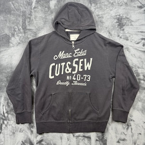 Op de afbeelding: Een antracietgrijze hoodie met rits, lange mouwen en twee voorzakken. Op de borst staat witte tekst gedrukt met de tekst Marc Ecko CUT & SEW No 40-73 Deadly Threads. Het kledingstuk is voorzien van een metalen ritssluiting over de gehele lengte.