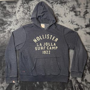 May include: A dark charcoal gray long-sleeve hooded sweatshirt featuring a front kangaroo pocket. The chest displays cream-colored text that reads HOLLISTER LA JOLLA SURF CAMP 1922. The garment has a hood with a drawstring-free neckline and ribbed cuffs.