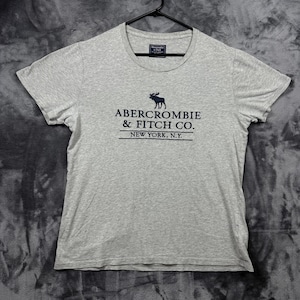 May include: A light grey short-sleeved t-shirt featuring a dark blue graphic of a moose silhouette above the text ABERCROMBIE & FITCH CO. NEW YORK, N.Y. The shirt has a crew neckline and is displayed on a black plastic hanger against a dark background.