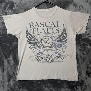 Puede incluir: Una camiseta de manga corta de color gris claro con un estampado gráfico azul oscuro en el pecho. El diseño incluye el texto "RASCAL FLATTS" sobre un logotipo circular con una "R" y alas, y una banda con "CHANGED TOUR".