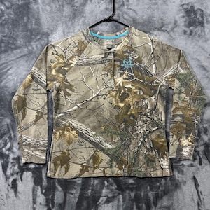 Könnte beinhalten: Ein langärmeliges Henley-Shirt mit einem Wald-Tarnmuster in Braun-, Beige- und Grüntönen. Das Shirt verfügt über eine Knopfleiste mit drei Knöpfen, einen Rundhalsausschnitt mit hellblauem Innenbesatz und das RealTree-Logo in Blau auf der Brust.