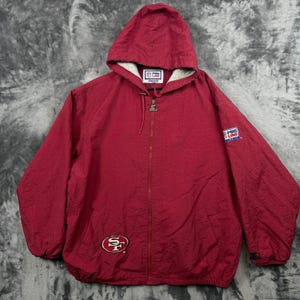 May include: A burgundy hooded windbreaker jacket featuring a full-length front zipper, long sleeves with elastic cuffs, and a drawstring hood. The jacket displays a San Francisco 49ers logo on the lower front and Pro Line Starter branding labels.