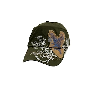May include: An olive green baseball cap featuring a white screen-printed floral scroll design on the front and side. A textured, tan-coloured heart-shaped patch with blue embroidered details is stitched onto the front panel of the cap.