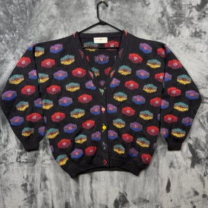 May include: A black V-neck cardigan sweater featuring a repeating pattern of multi-colored, textured circular shapes in red, blue, yellow, and green. The garment has long sleeves, a ribbed hem and cuffs, and a front button closure with colorful buttons.