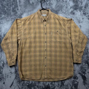May include: A long-sleeve button-down shirt in a tan and brown plaid pattern. The shirt features a button-down collar, a single chest pocket with a small Columbia brand tag, and a buttoned front placket. The fabric has a textured appearance.