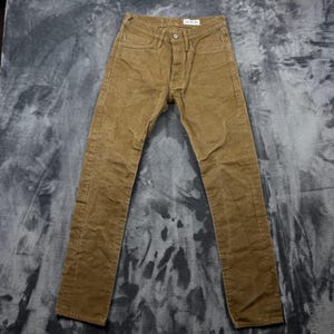 May include: A pair of brown pants featuring a button fly closure, five pockets, and a visible brand label on the interior waistband. The pants have a straight leg cut with visible stitching along the seams and a textured fabric finish throughout.