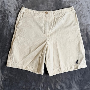 Könnte beinhalten: Beige Shorts aus Baumwolle mit einem Bund mit Gürtelschlaufen, einem einzelnen Knopfverschluss und einem Reißverschluss. Die Shorts verfügen über zwei Seitentaschen und ein kleines, dunkelblau gesticktes Mickey-Mouse-Logo am rechten Bein.