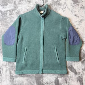 Op de afbeelding: Een saliegroene fleecejas met een volledige ritssluiting aan de voorzijde, een hoge kraag en twee verticale zijzakken. De mouwen zijn voorzien van contrasterende donkerblauwe ovale elleboogstukken en de zoom en boorden hebben een textuur.