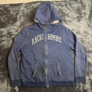 May include: A blue zip-up hoodie featuring a cream-colored sherpa lining inside the hood. The front displays the text ABERCROMBIE in white lettering across the chest. The garment includes a full-length front zipper and two front pockets.
