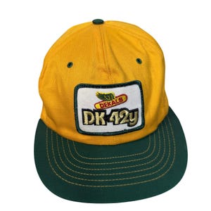 Puede incluir: Una gorra de béisbol dorada con visera verde oscuro y un parche rectangular central. El parche presenta un logotipo alado verde sobre el texto rojo DEKALB y el texto grande en negro DK 42y. La gorra incluye ojales de color verde oscuro.