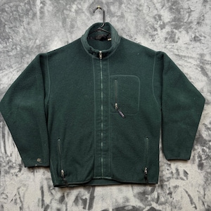 May include: A dark forest green fleece jacket featuring a full-length front zipper, a high mock neck collar, and long sleeves. The garment includes one vertical zippered chest pocket on the left side and two lower vertical zippered pockets.