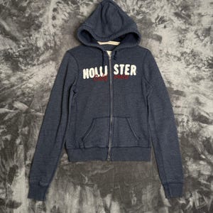 May include: A dark blue zip-up hoodie featuring a hood with drawstrings, long sleeves, and two front pockets. The chest displays the white text HOLLISTER with the red cursive word California underneath. The garment has a full-length metal zipper.