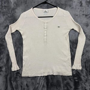 May include: A long-sleeve, cream-colored henley shirt featuring a ribbed texture, a round neckline, and a five-button placket. The garment includes a small green embroidered crocodile logo on the left chest and a Lacoste brand label at the neck.