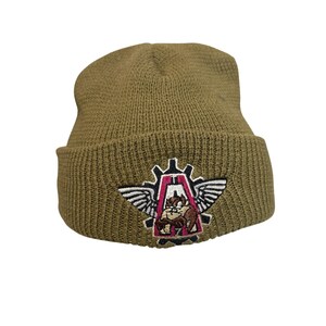 Puede incluir: Un gorro de punto color verde oliva con borde doblado y un parche bordado en la parte frontal. El parche muestra una forma de engranaje estilizada con alas blancas y un emblema central rosa y negro que representa un personaje animado marrón.
