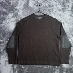 May include: A dark brown long-sleeve sweater featuring a crew neckline and contrasting grey elbow patches on both sleeves. The garment has a ribbed knit texture at the cuffs and hem, and it is displayed on a black plastic hanger.