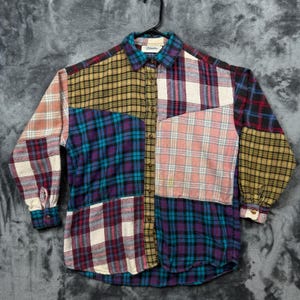 May include: A long-sleeve button-down shirt featuring a patchwork design of various plaid patterns in colours including mustard yellow, teal, purple, pink, white, and black. The shirt has a standard collar and a central button placket.