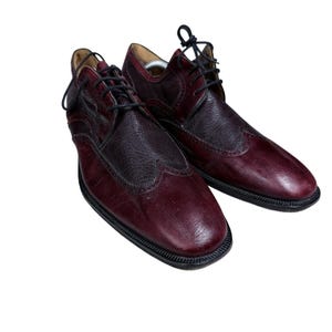 May include: A pair of burgundy and dark brown leather oxford shoes featuring a wingtip design with textured leather panels, black laces, and black soles. The shoes have a polished finish and a rounded toe, showcasing a two-tone color scheme.