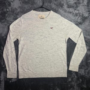 May include: A light gray heathered long-sleeve sweater with a crew neckline and ribbed cuffs and hem. The garment features a small embroidered dark red bird logo on the left chest and a Hollister brand label inside the collar.