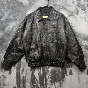 May include: A black leather jacket featuring a collared neckline, long sleeves, and a front button closure. The jacket is constructed from multiple stitched panels of leather, creating a textured surface, and includes two side pockets and a ribbed hem.