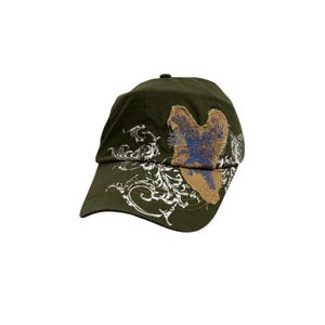 May include: An olive green baseball cap featuring a white screen-printed floral scroll design on the front and side. A textured, tan-coloured heart-shaped patch with blue embroidered details is stitched onto the front panel of the cap.