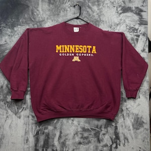 Puede incluir: Una sudadera granate de manga larga con cuello redondo que presenta el texto MINNESOTA GOLDEN GOPHERS en letras amarillas en el pecho, junto con un logotipo M amarillo. La prenda se muestra en una percha de plástico negra sobre un fondo gris.