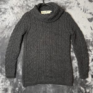 Könnte beinhalten: Ein dunkelgrauer Langarmpullover mit einem markanten Wasserfallkragen und einem durchgehenden vertikalen Zopfstrickmuster. Das Kleidungsstück verfügt über ein weißes Innenetikett am Hals mit dem in Schwarz gedruckten Text ARAN MÓR.