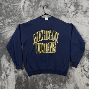 May include: A navy blue long-sleeve crewneck sweatshirt featuring the text MICHIGAN WOLVERINES printed in yellow with a thin white outline across the chest. The garment is displayed on a black plastic hanger against a textured grey background.
