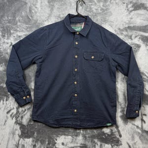 Peut inclure: Une chemise à manches longues bleu marine avec un col pointu, une poche poitrine boutonnée et des poignets boutonnés. La doublure intérieure du col présente un motif à carreaux rouge, blanc et vert avec une étiquette de marque Woolrich.