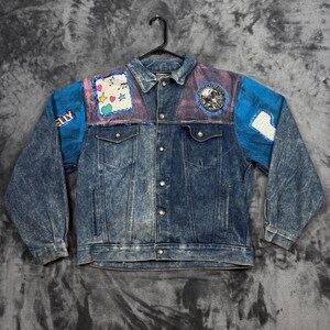 May include: A blue acid-wash denim jacket featuring a button-up front, two chest pockets, and two side pockets. The upper chest area includes a white patch with musical notes and colourful stars and hearts, plus a circular patch with a graphic design.