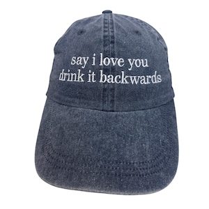 May include: A dark blue denim baseball cap with a six-panel construction and a curved brim. The front of the cap features white embroidered text that reads, "say i love you drink it backwards" in a simple, lowercase serif font style.